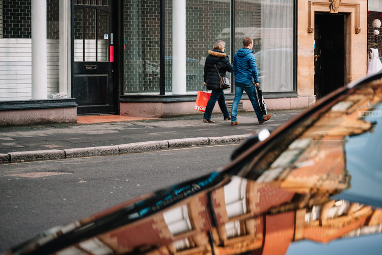 People walking on Howard Street, with buildings reflected in the parked car in the foreground